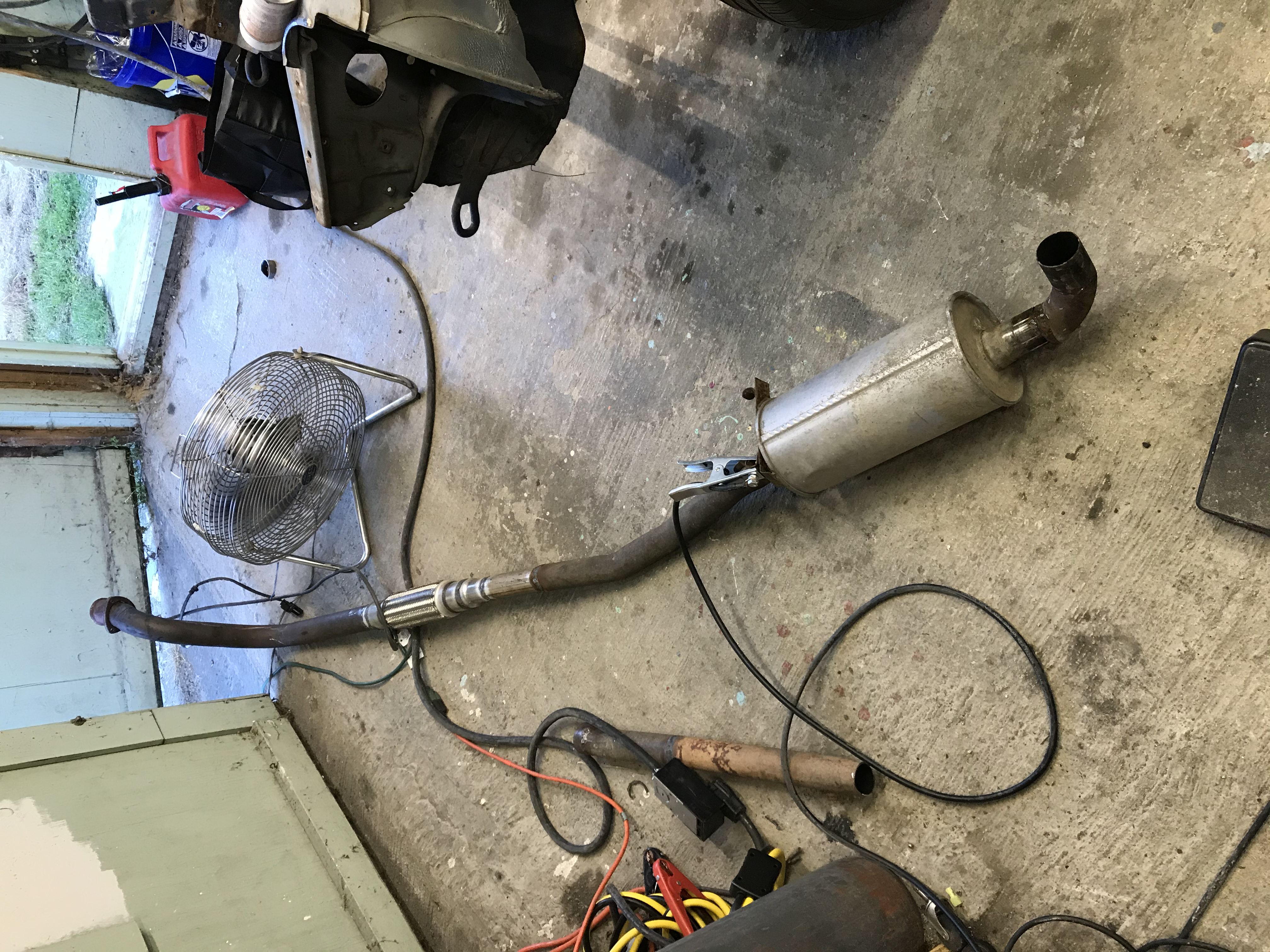 Metal exhaust tubing with a single muffler on a concrete floor with a wire fan and a welding electrode clamped to the tubing
