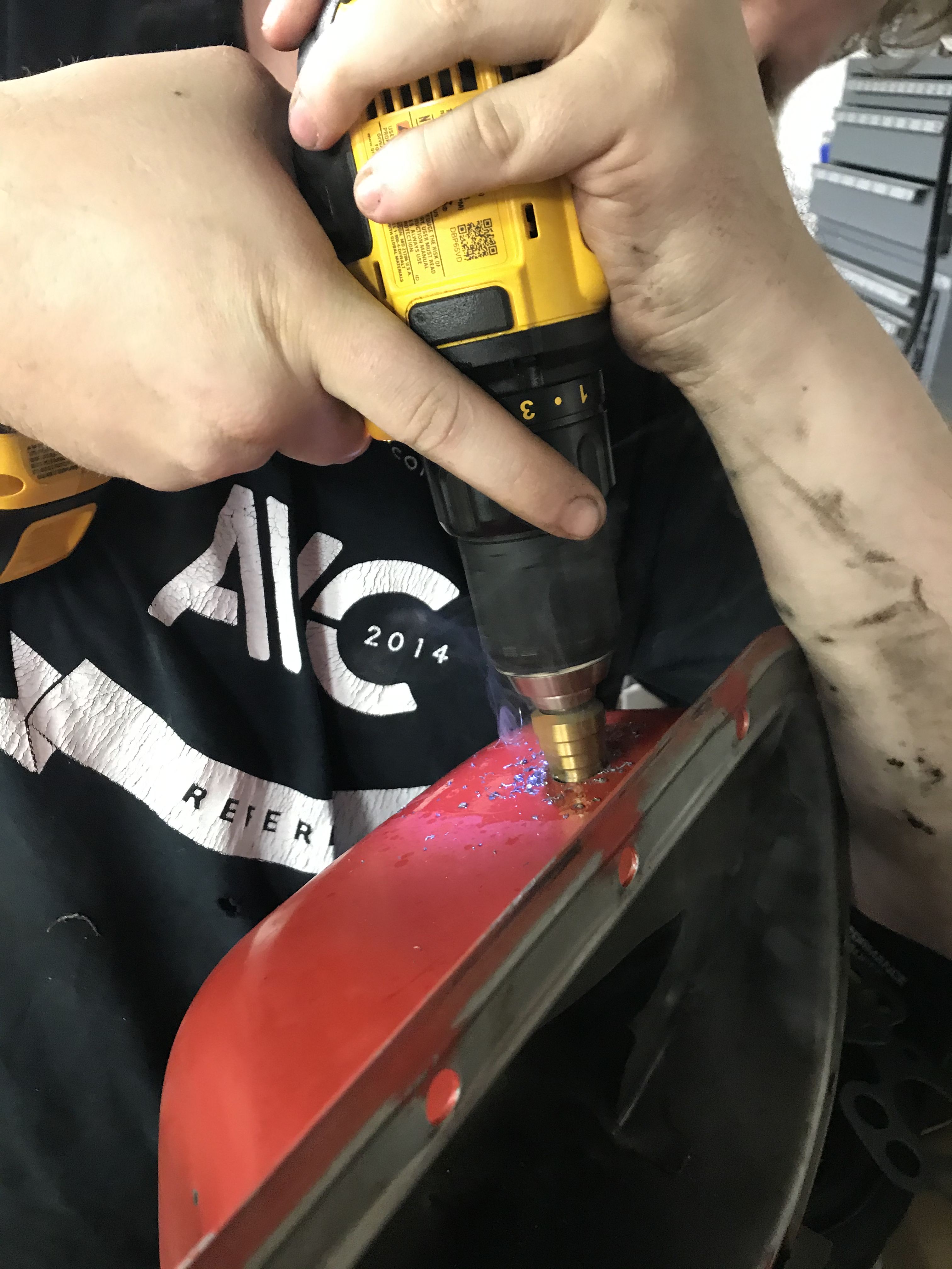 Man using a cordless drill with a brass step bit to drill into a metal oil pan
